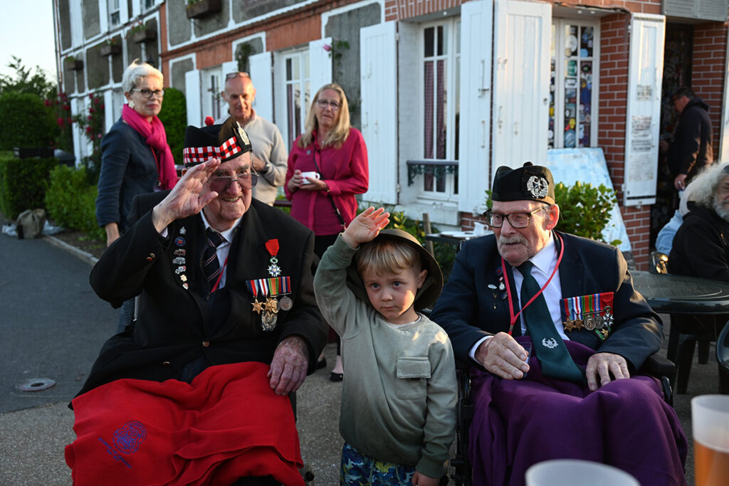5 years old boy wearing a tin helmet salutes with 2 veterans